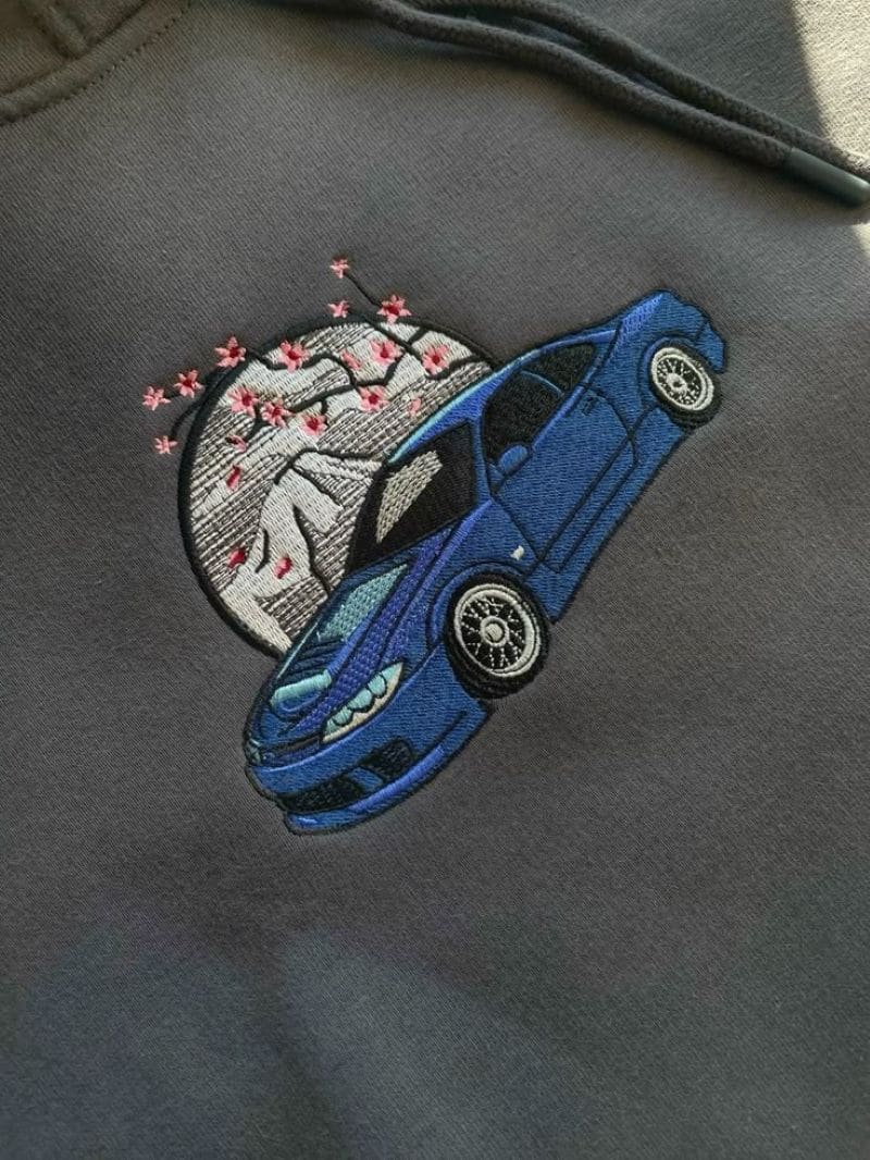 Gesticktes blaues Sportauto vor Kirschblüten-Mond-Motiv auf grauem Hoodie